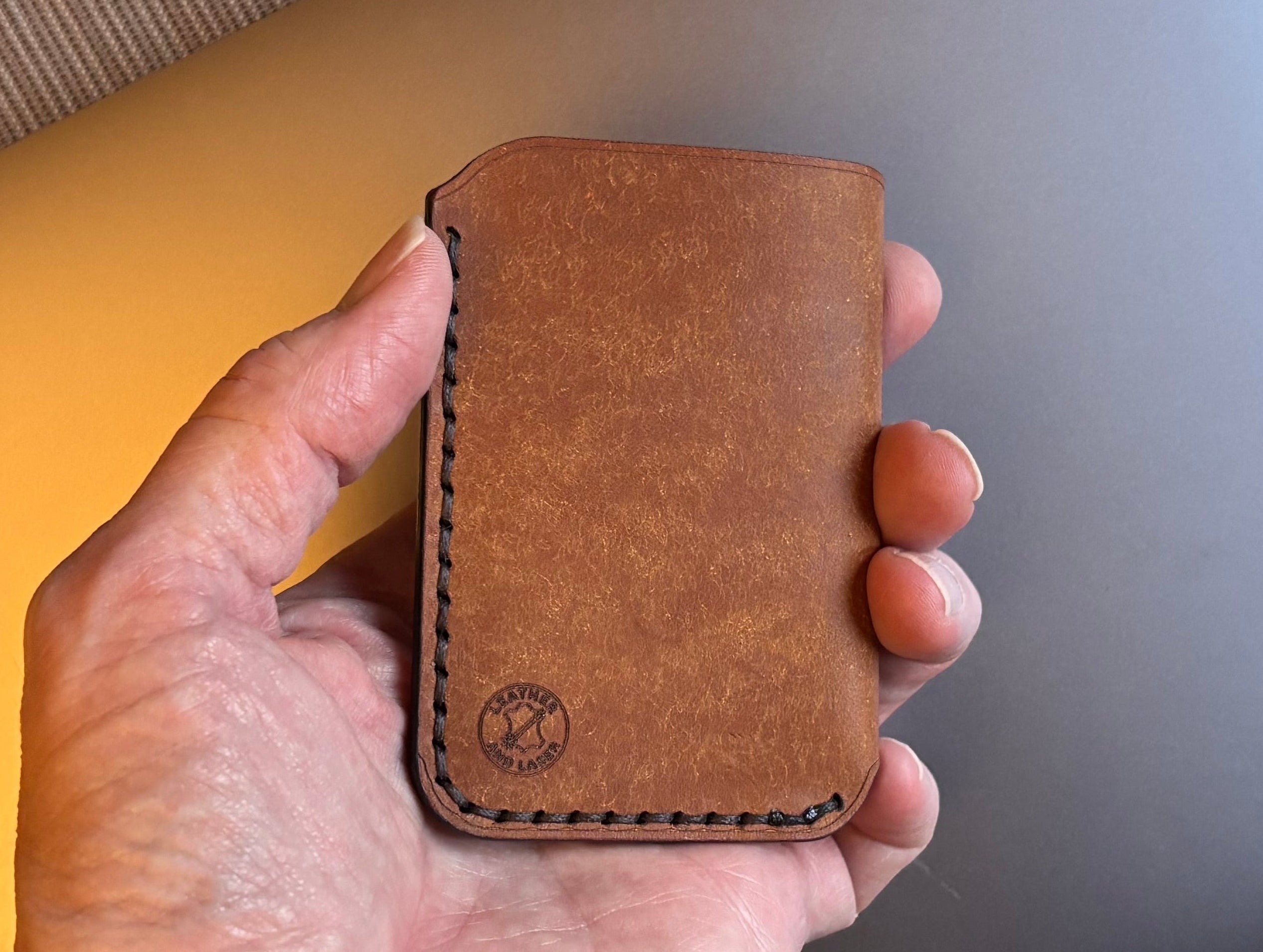 Hand holding a brown leather cardholder with a laptop in the background